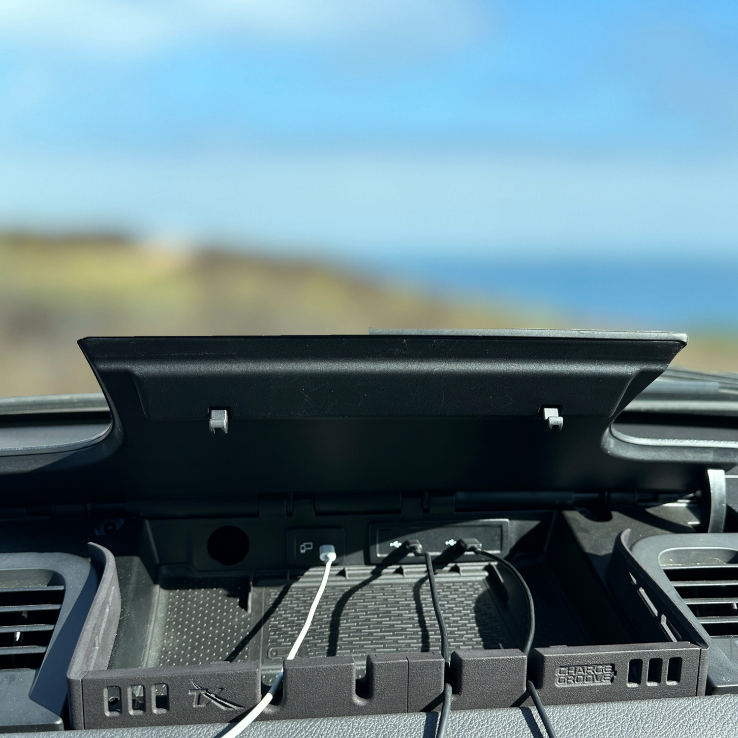 Car dashboard organizer with charging cables plugged in, lid open against a scenic background.