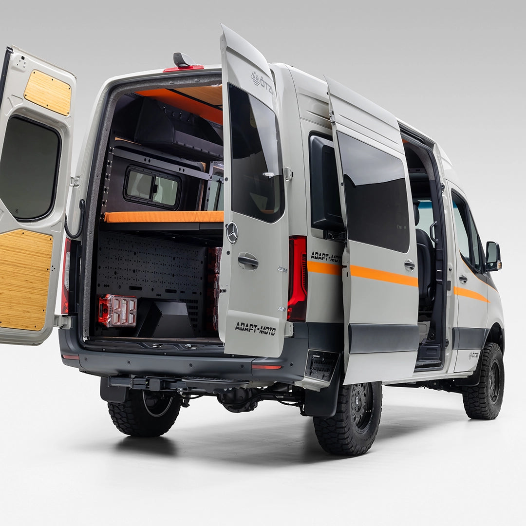 Cozy off‑road camper van interior with raised bed, wood paneling, and mountain views through rear doors
