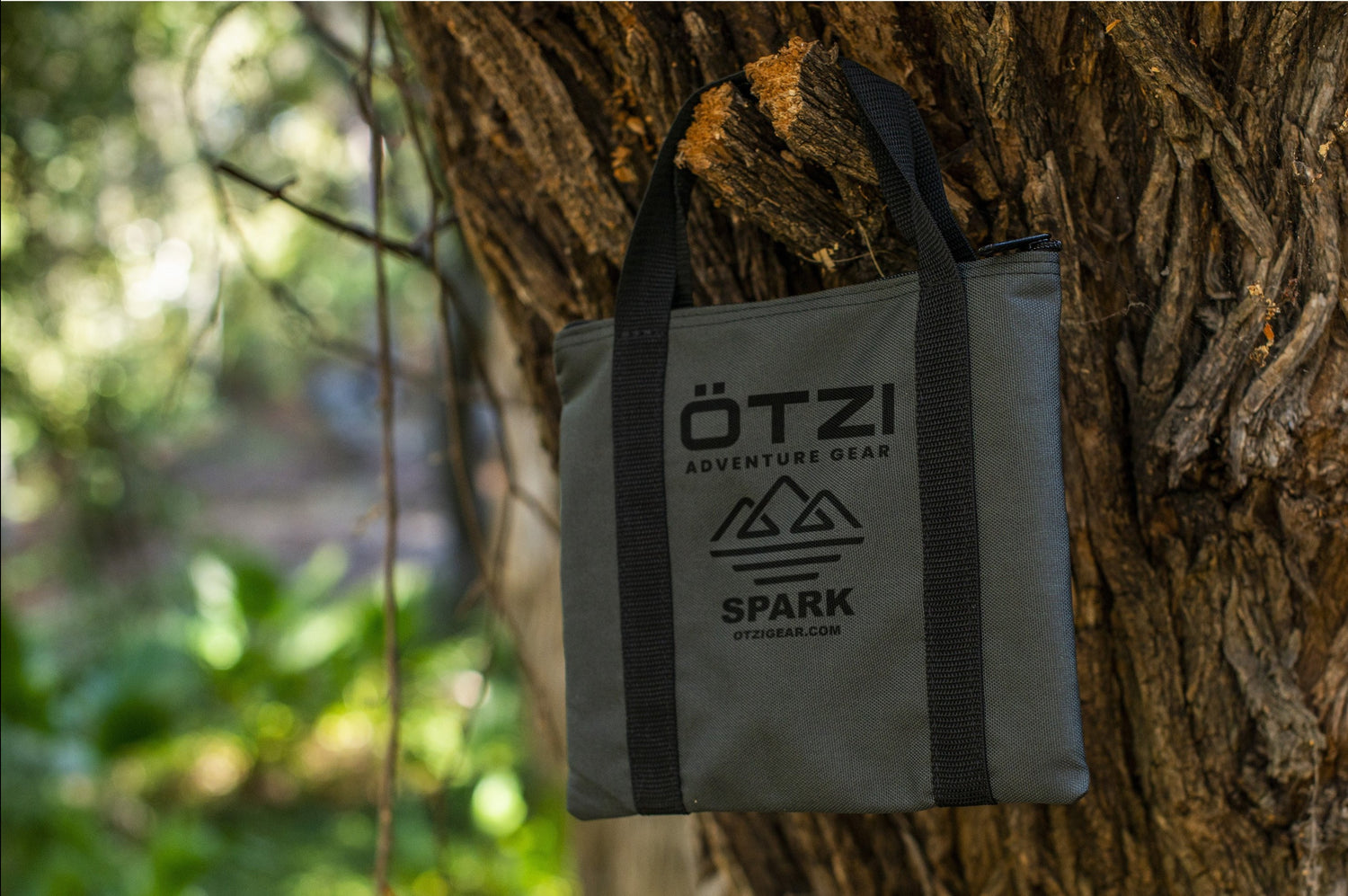 Gray Otzi Spark gear bag with black handles hanging on a tree trunk outdoors.