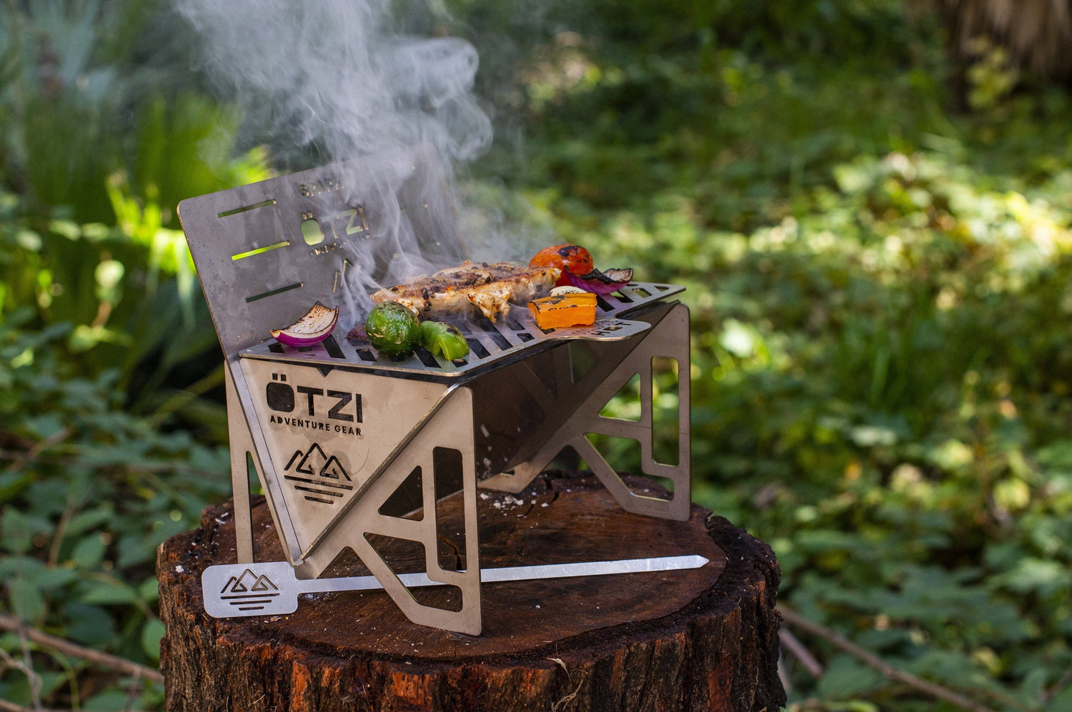 Portable stainless steel grill cooking meat and vegetables in an outdoor forest setting.