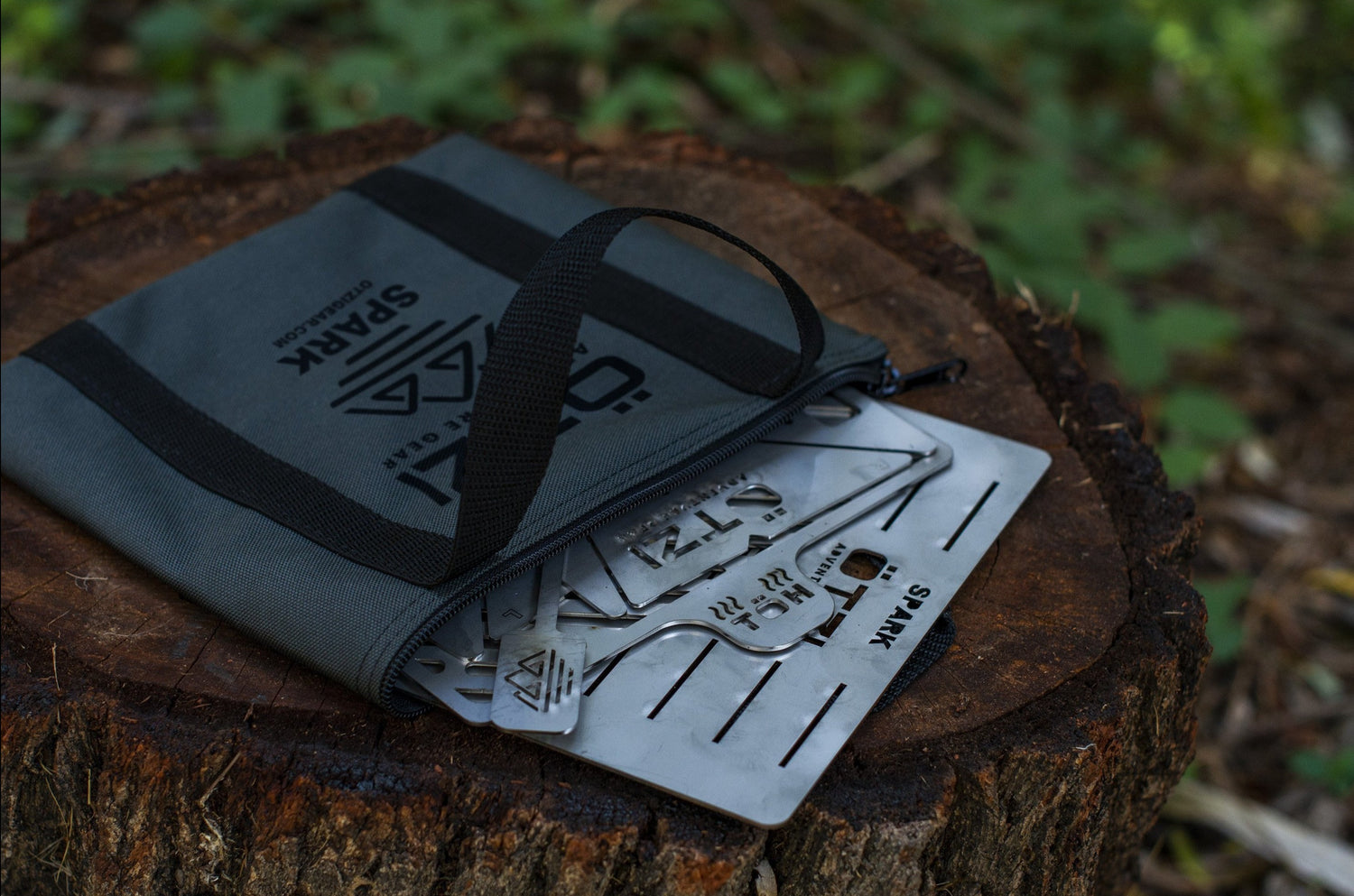 Metal camping stove kit partially pulled from a gray zippered pouch on a tree stump outdoors