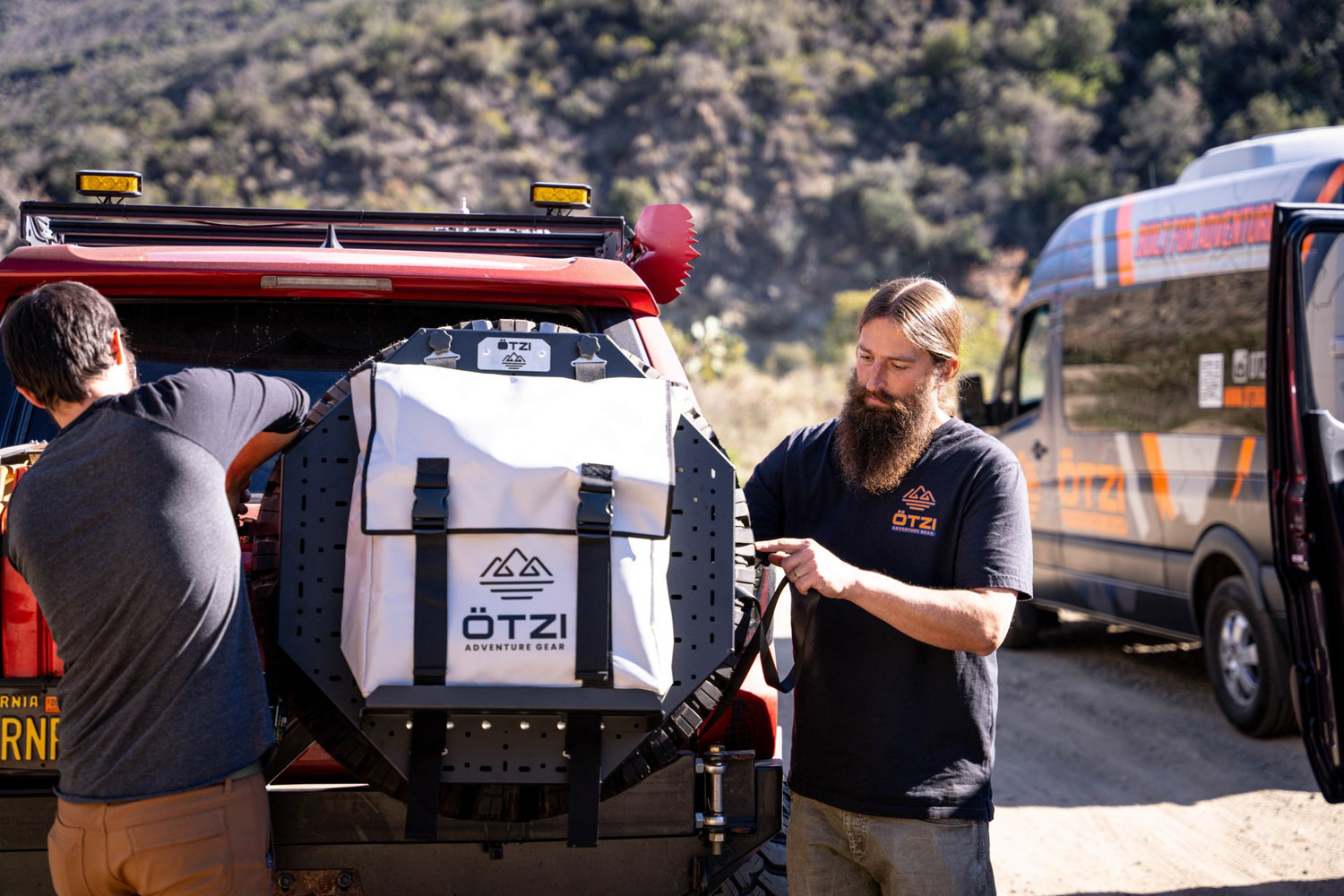 Two men installing a white Ötzi gear bag on the spare tire of an off-road vehicle