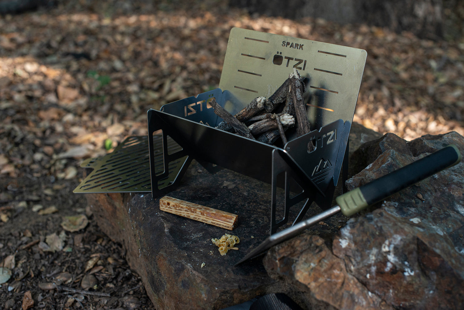 Compact metal campfire stove with wood pieces, set on rocks in a forest setting