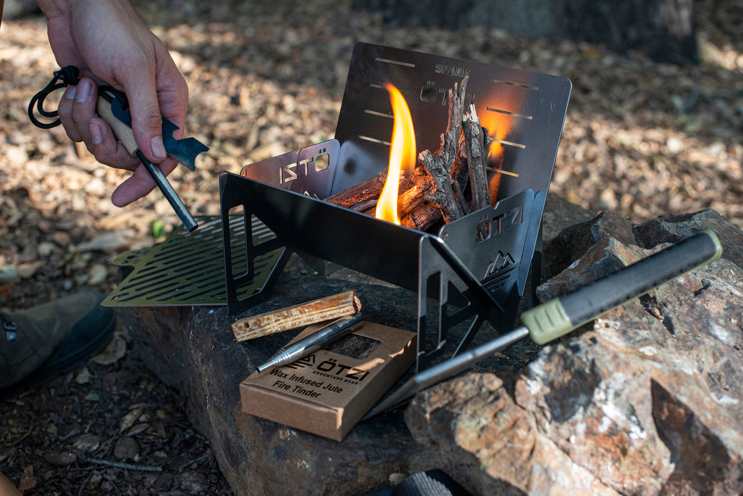Portable metal camping stove with kindling and lit fire, placed on a rock in a forest setting.