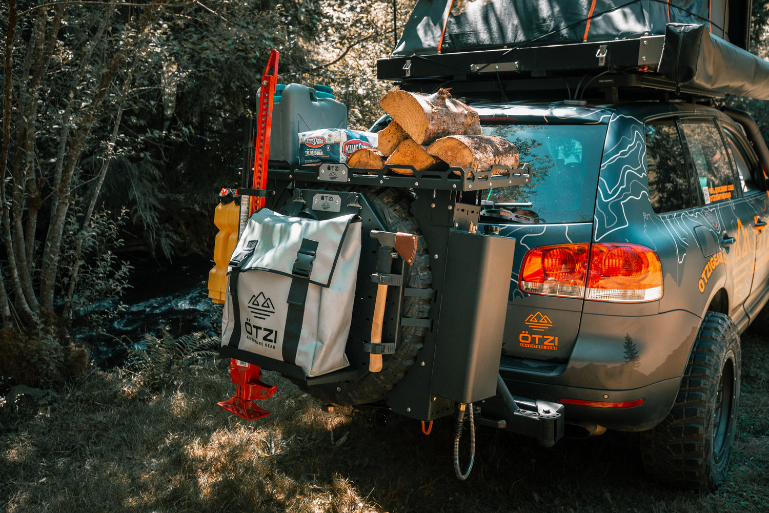 Off-road SUV with rear cargo rack carrying firewood, gear bag, and tools in a forest setting