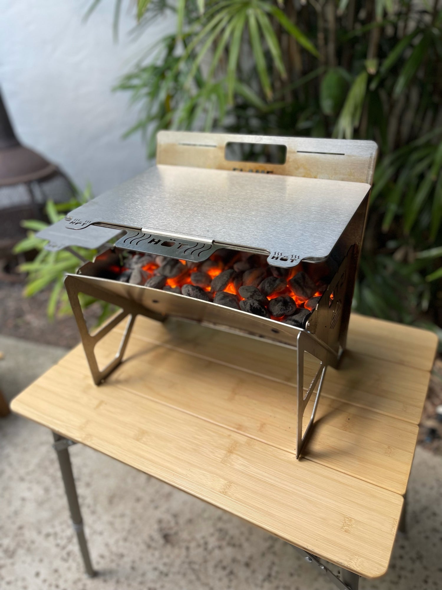 Portable stainless steel charcoal grill with glowing coals, set on a wooden table outdoors.