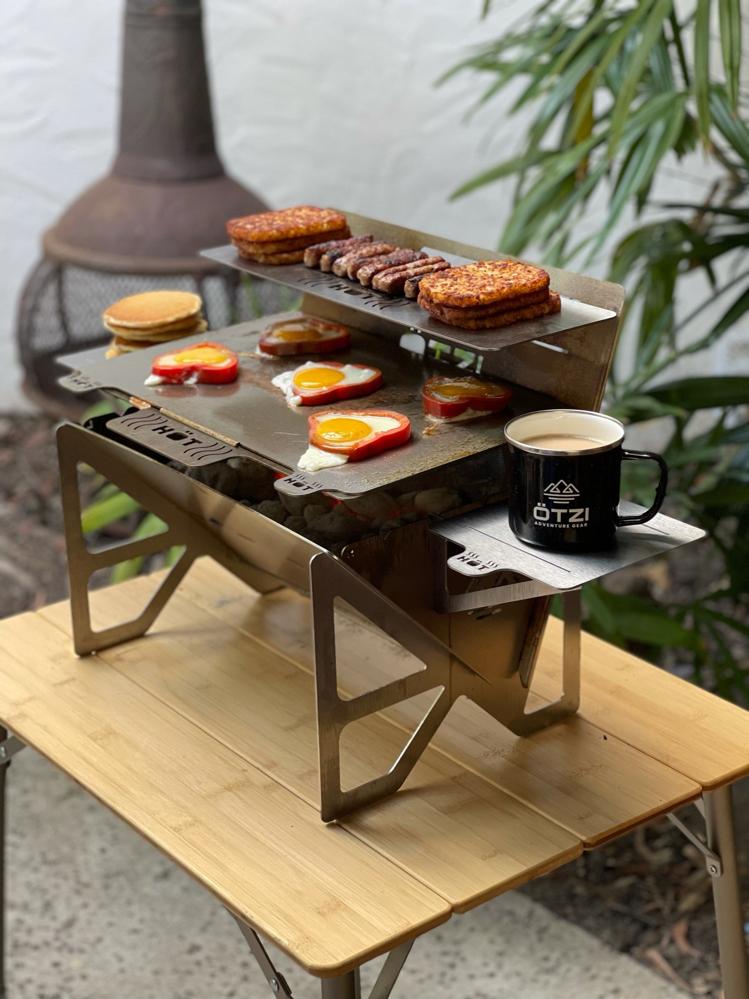 Portable stainless steel grill cooking burgers, eggs, and sausage outdoors on a wooden table.