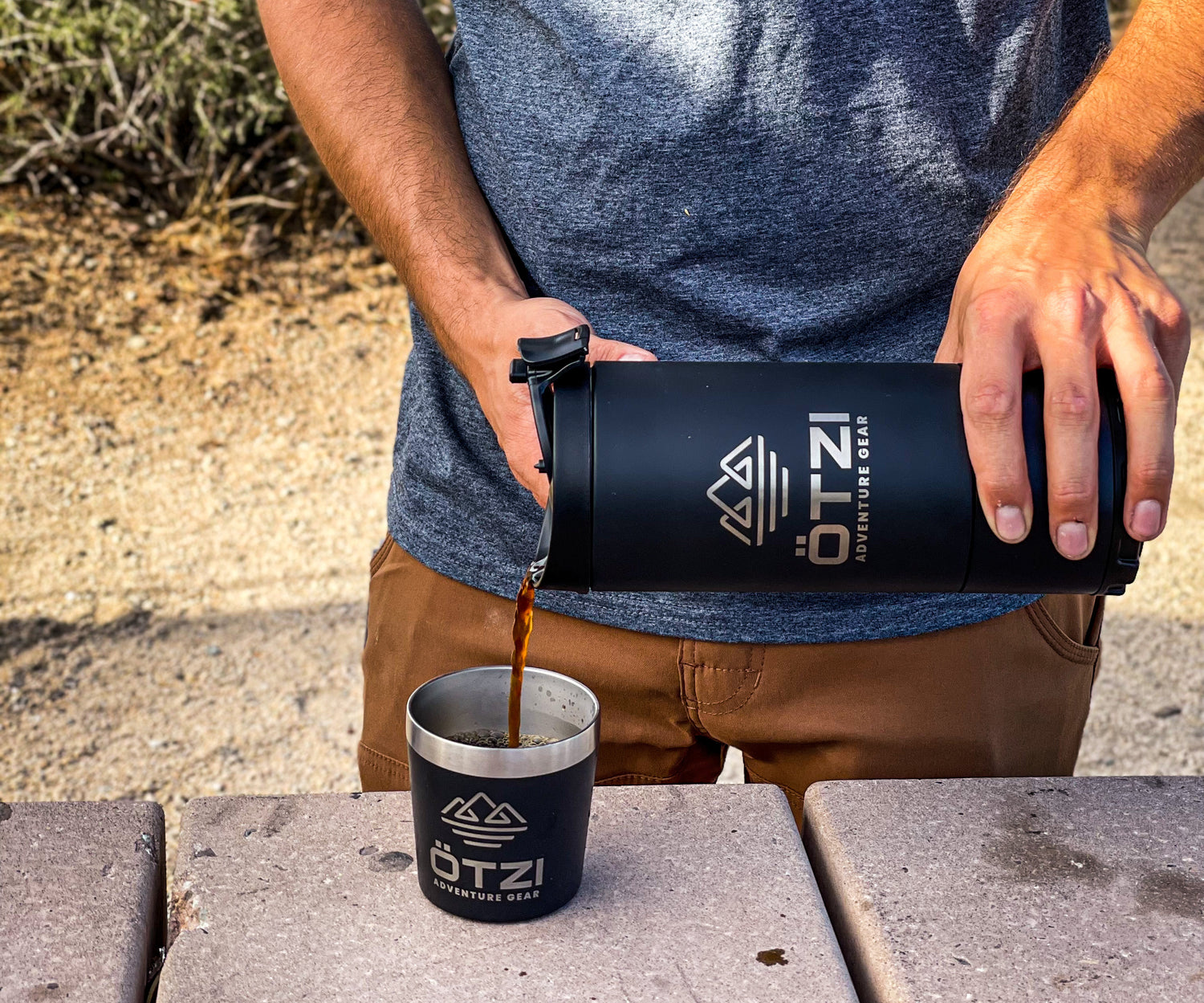 Person pouring coffee from a black insulated bottle into a matching metal cup outdoors.