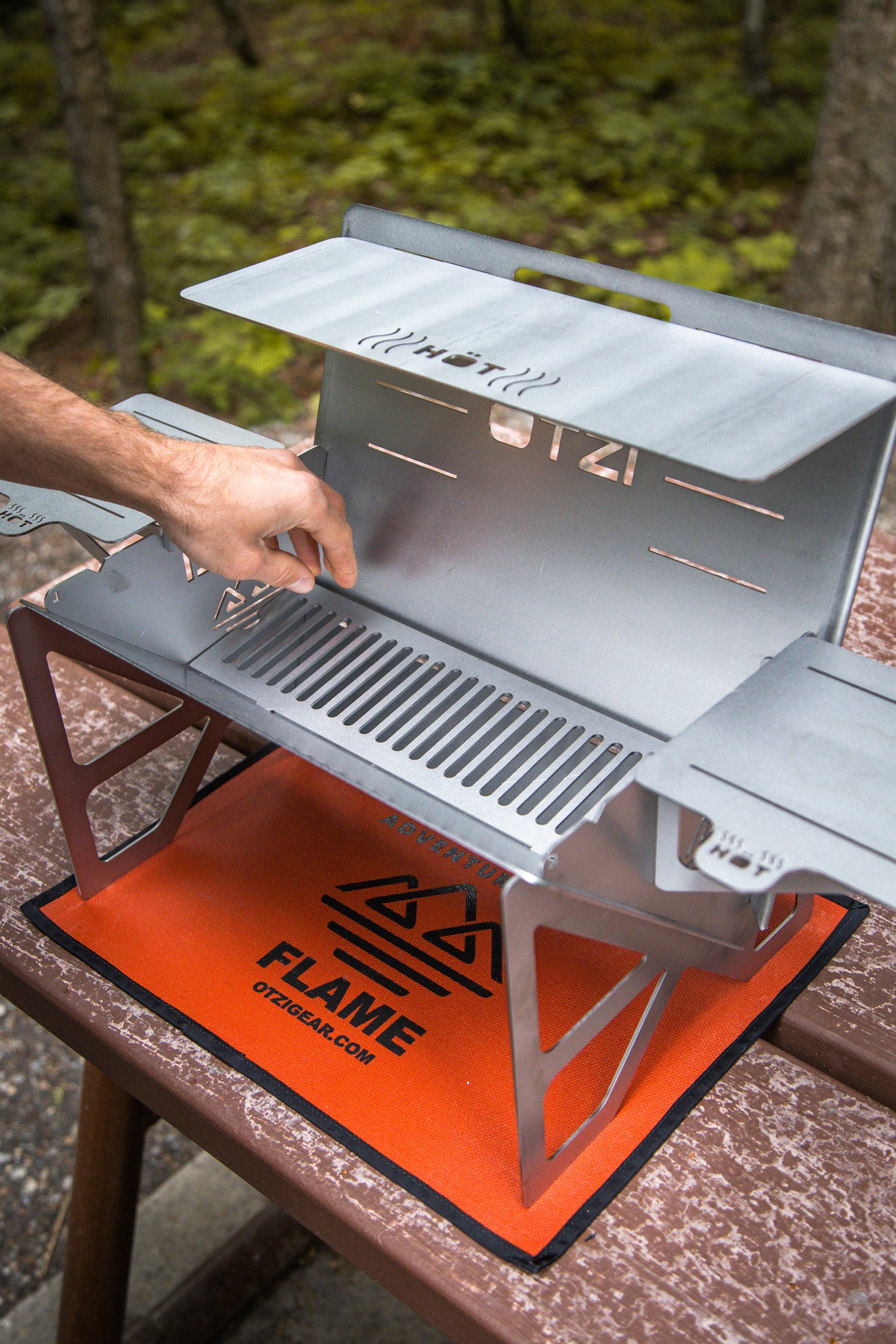 Person opening a portable metal camping grill on a picnic table in a forest setting
