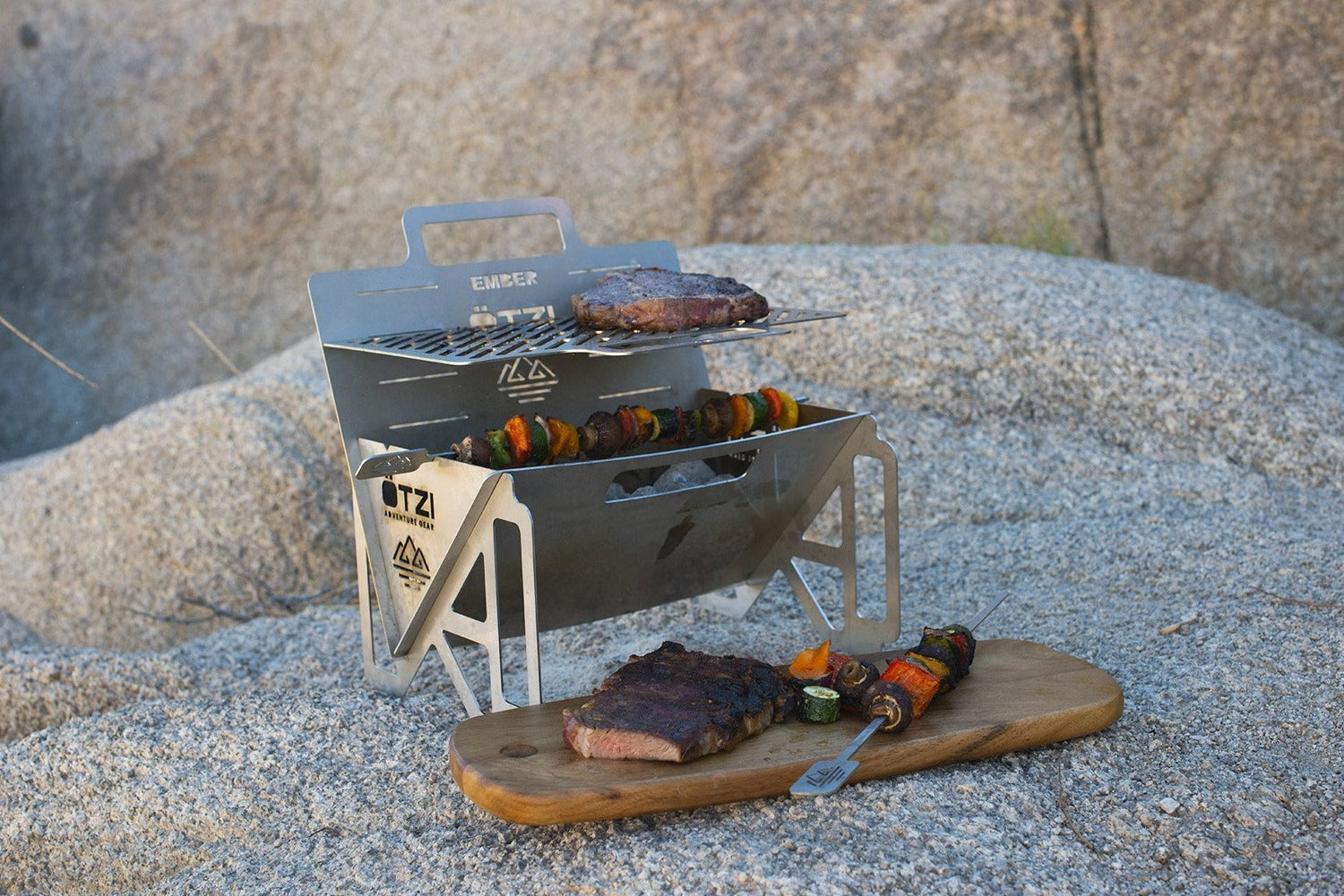 Portable stainless steel grill with steak and skewers, set on a sandy outdoor surface.
