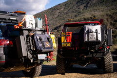 Two off-road vehicles with mounted gear bags, fuel cans, and recovery tools in a mountain setting.
