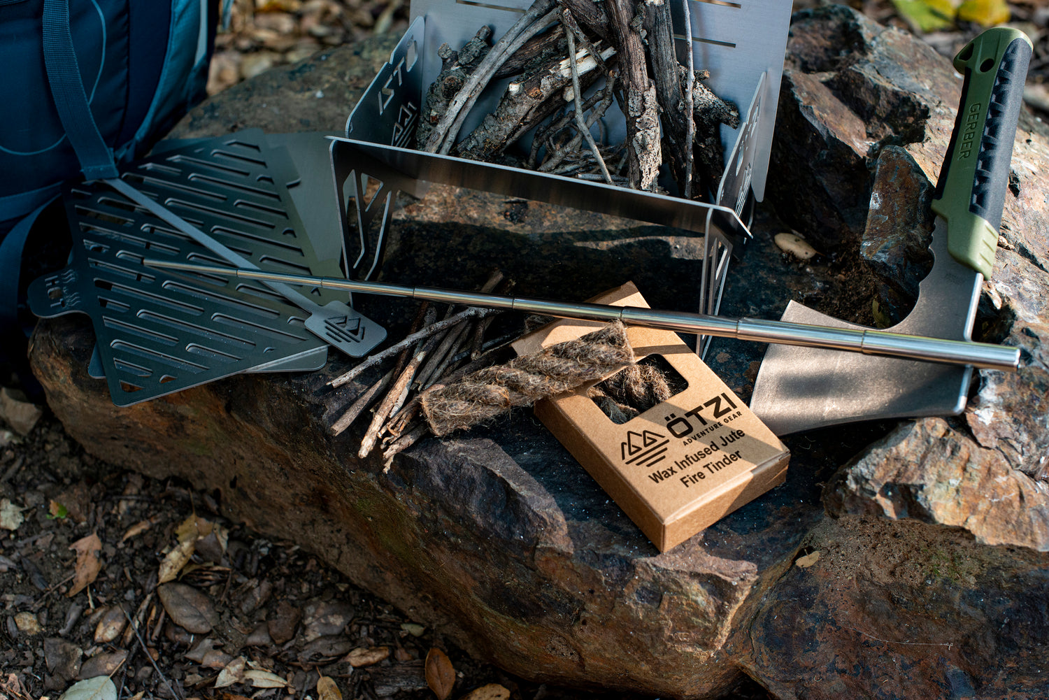 Portable stainless steel campfire grill set with skewers and fire starter on a rock in the woods.