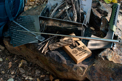 Portable stainless steel campfire grill set with skewers and fire starter on a rock in the woods.