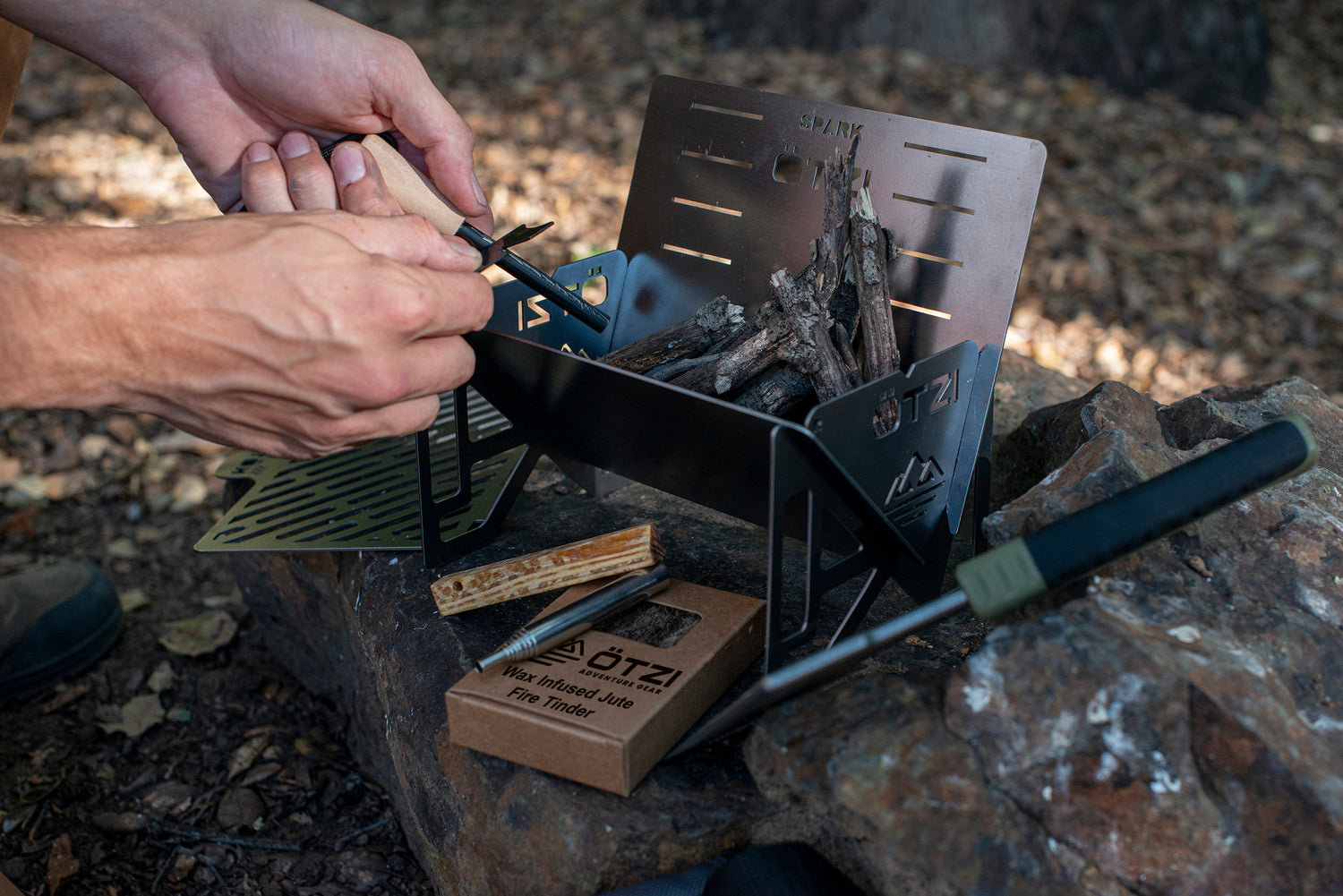 Hands preparing a compact outdoor fire pit with kindling and a fire starter in a forest setting.