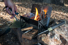 Portable metal camping stove with kindling and lit fire, placed on a rock in a forest setting.