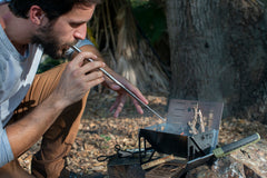 Man using fire blower to ignite kindling in a portable metal campfire pit outdoors.
