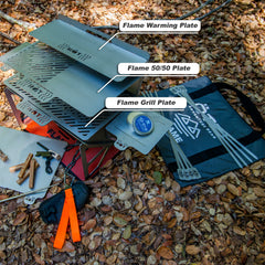 Portable outdoor grill with labeled warming, 50/50, and grill plates on forest ground with accessories.