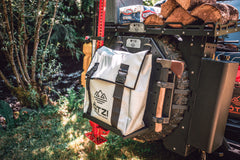 Overland gear mounted on a vehicle’s spare tire including a white storage bag, axe, and firewood.