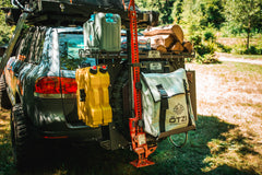 SUV rear with mounted gear, including water cans, a recovery jack, firewood, and an ÖTZI storage bag.