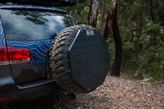 Mounted spare tire with black protective cover on SUV parked in wooded area