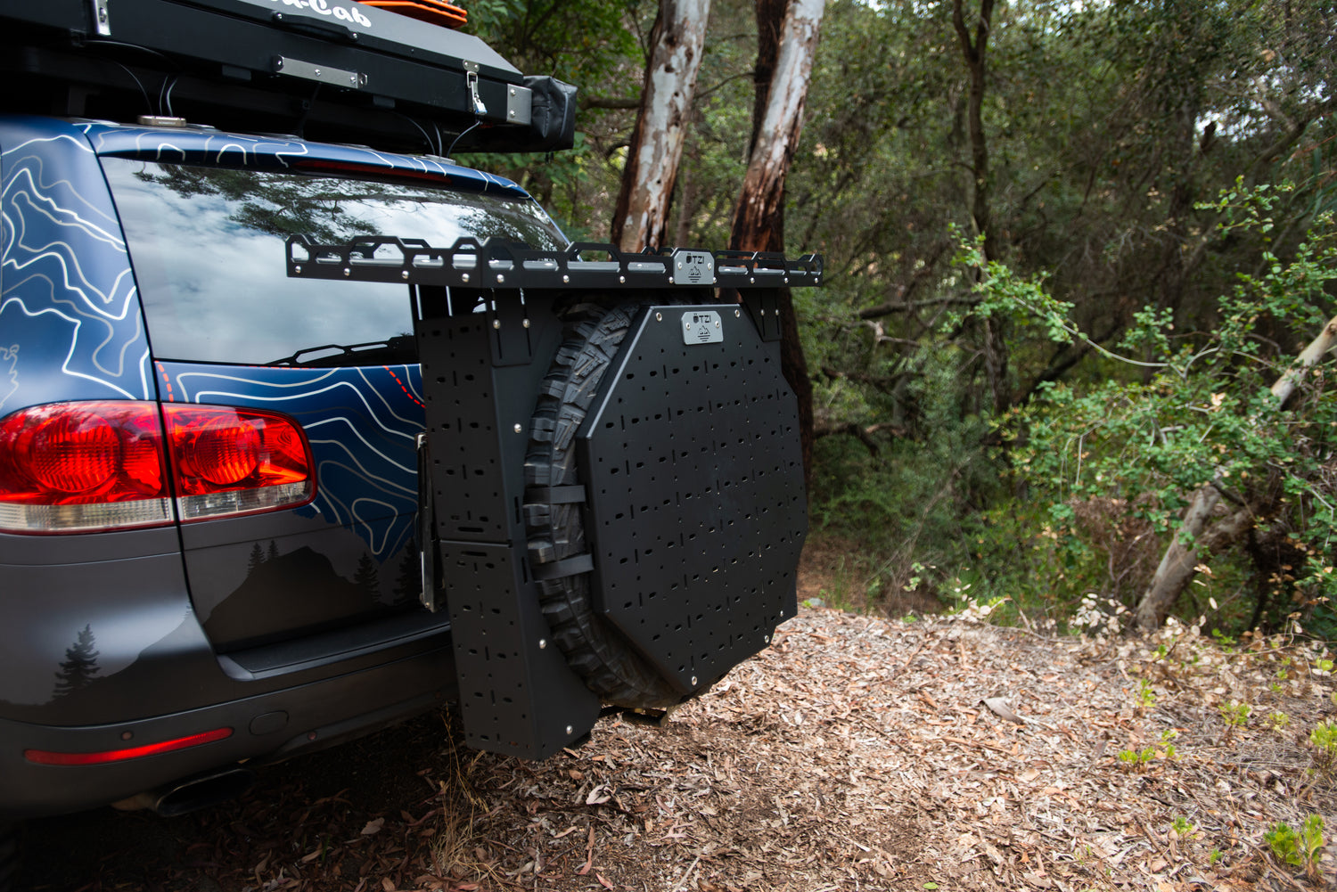 Off-road vehicle with swing-out tire carrier and mounted gear panel in a wooded area
