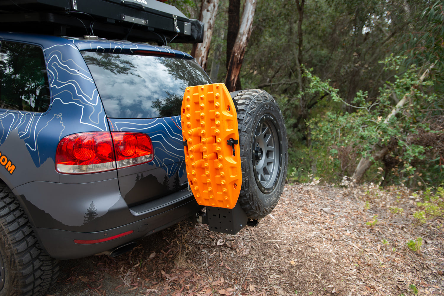 Orange traction board mounted on the rear tire carrier of an off-road SUV in a forest setting.