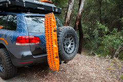 Orange recovery traction board mounted on the rear swing-out of an off-road SUV in a wooded area.
