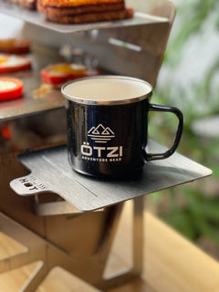Black OTZI mug on a metal camping grill shelf with food in the background