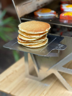 Stack of golden pancakes on a metal tray beside a griddle with eggs cooking