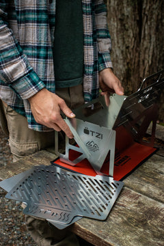 Person assembling a stainless steel portable camping grill on a picnic table outdoors.