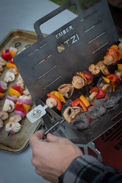 Person grilling skewers with vegetables and meat on a portable metal grill outdoors.