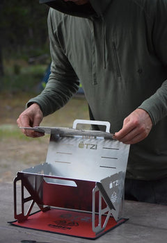 Person assembling a stainless steel portable camping stove on a picnic table.