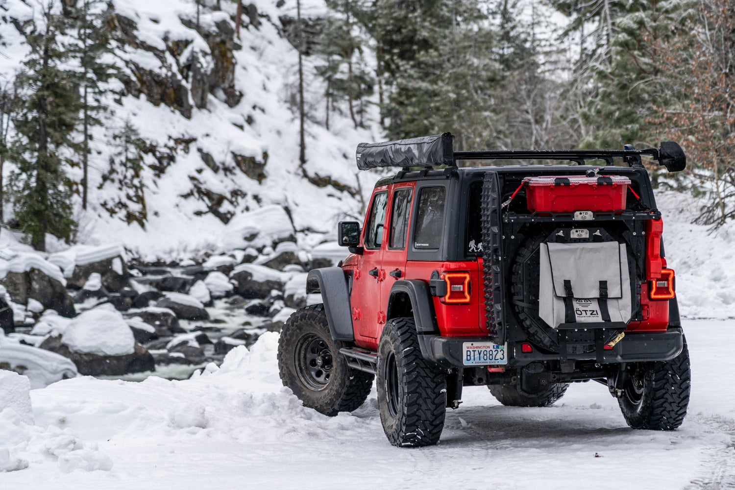 Red off-road SUV with storage gear parked on snowy trail near a river in winter forest