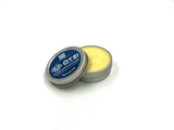 Open tin of Ö.TZI balm with yellow salve inside, lid resting against container on white background.